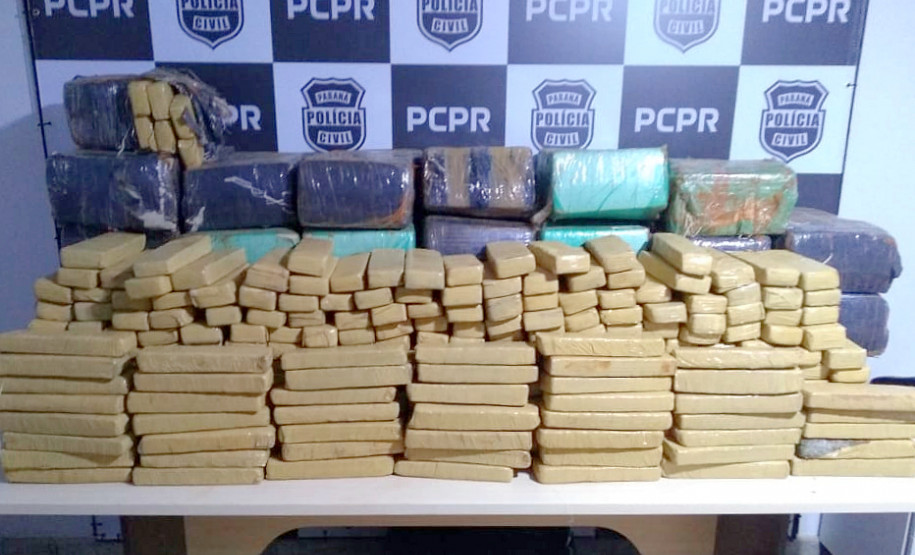 Drogas apreendidas empilhadas sobre uma mesa. Ao fundo, banner da Polícia Civil