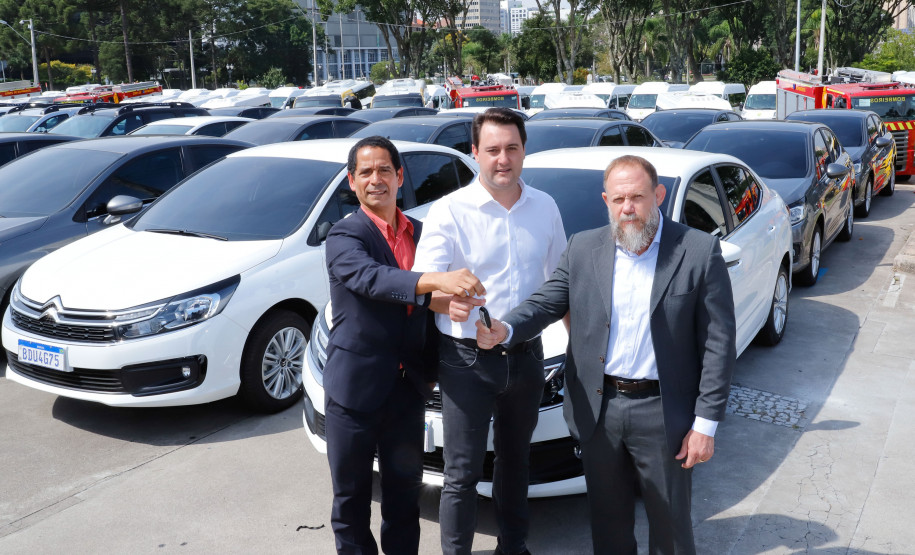 Secretário de Segurança Pública, Governador e Delegado Geral posando para foto. Ao fundo, veículos entregues pelo Governo.