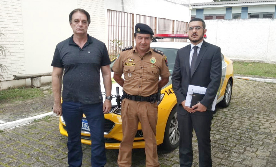 Participantes da reunião em pé, posando para foto. Ao fundo, viatura da polícia militar