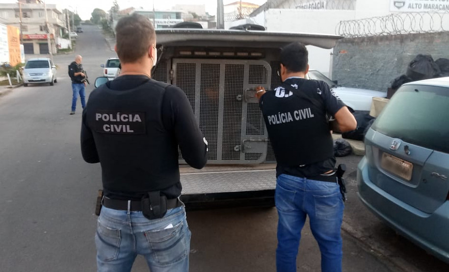 Policiais civis em pé, ao lado de viatura com suspeito em seu interior