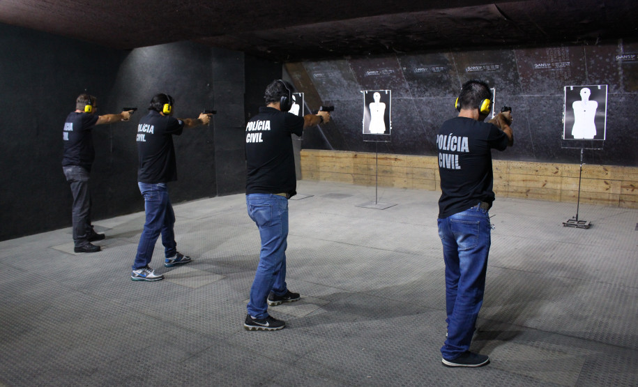 Policiais civis alinhados em pé, apontando suas armas para alvos de papel, no interior de um estande de tiro