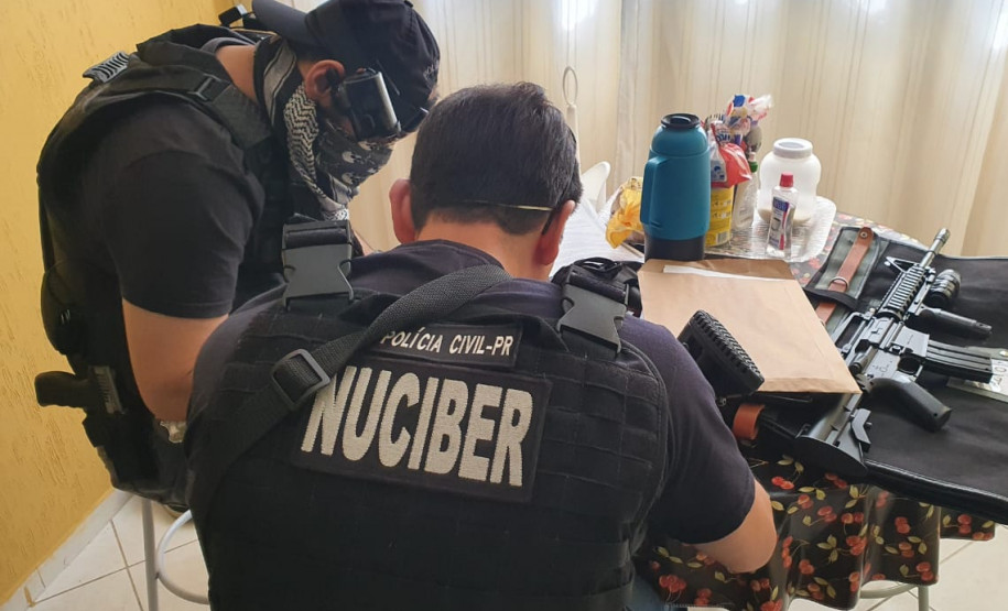 Dois policiais civis sentados, com objetos apreendidos sobre uma mesa.