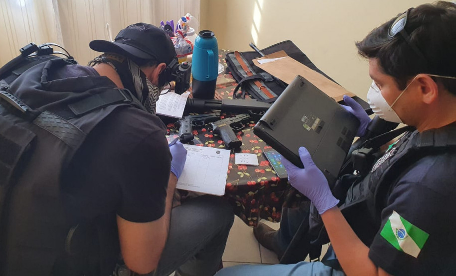 Dois policiais civis sentados, com objetos apreendidos sobre uma mesa.