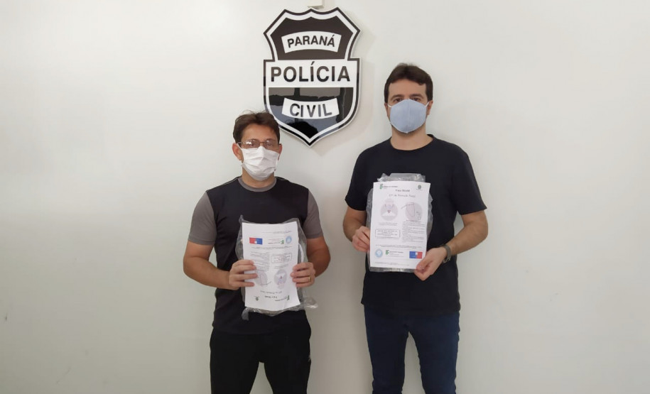 Policiais civis segurando embalagens com as máscaras. Ao fundo, brasão da Polícia Civil.