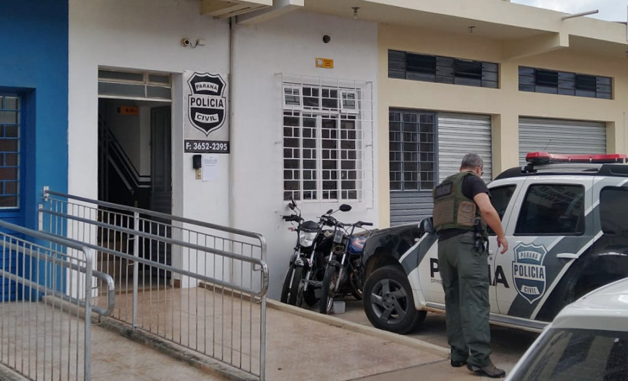 Policial civil ao lado de viatura, em frente à delegacia