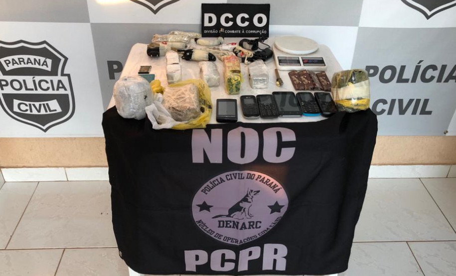 Materiais apreendidos, sobre uma mesa. Ao fundo, banner da polícia civil