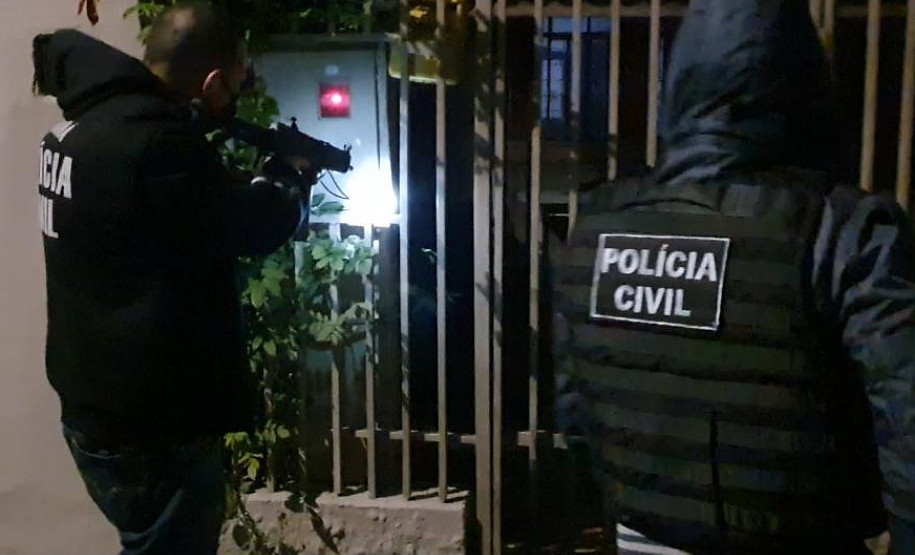 Dois policiais civis em frente a um portão. Um deles aponta uma arma.
