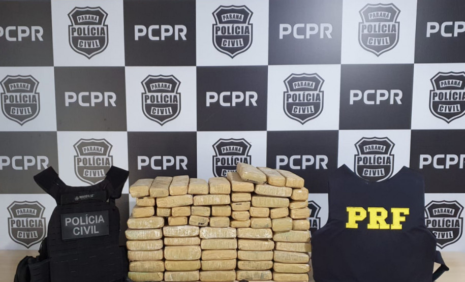 Diversos tabletes de droga entre coletes da polícia civil e policia rodoviária federal. ao fundo, banner da polícia civil