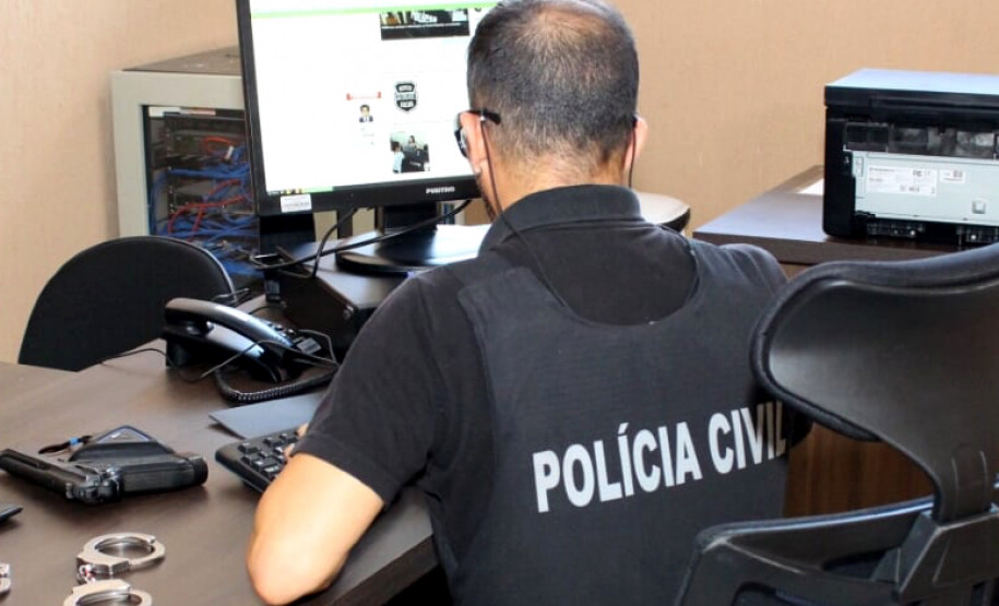 Policial ao computador. Na mesa, arma e algemas