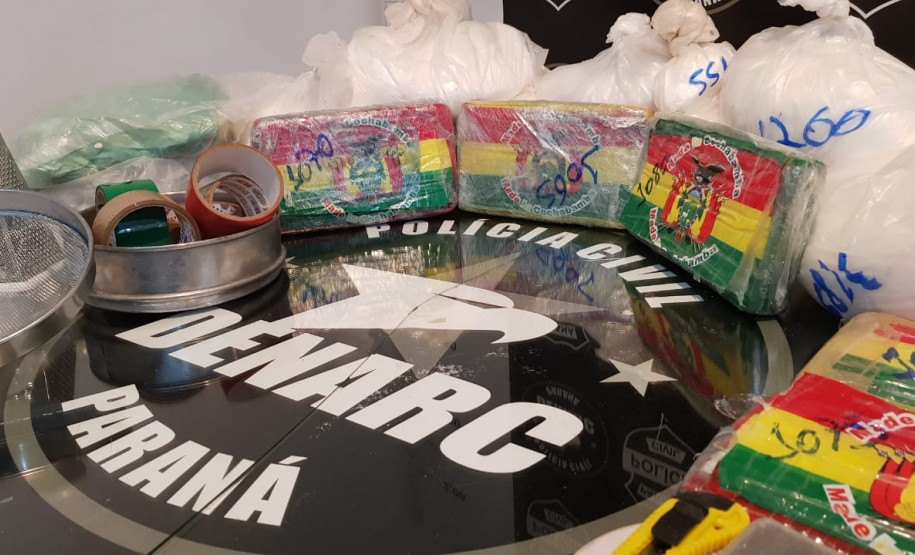 Material apreendido, sobre uma mesa. Ao centro, logomarca da DENARC