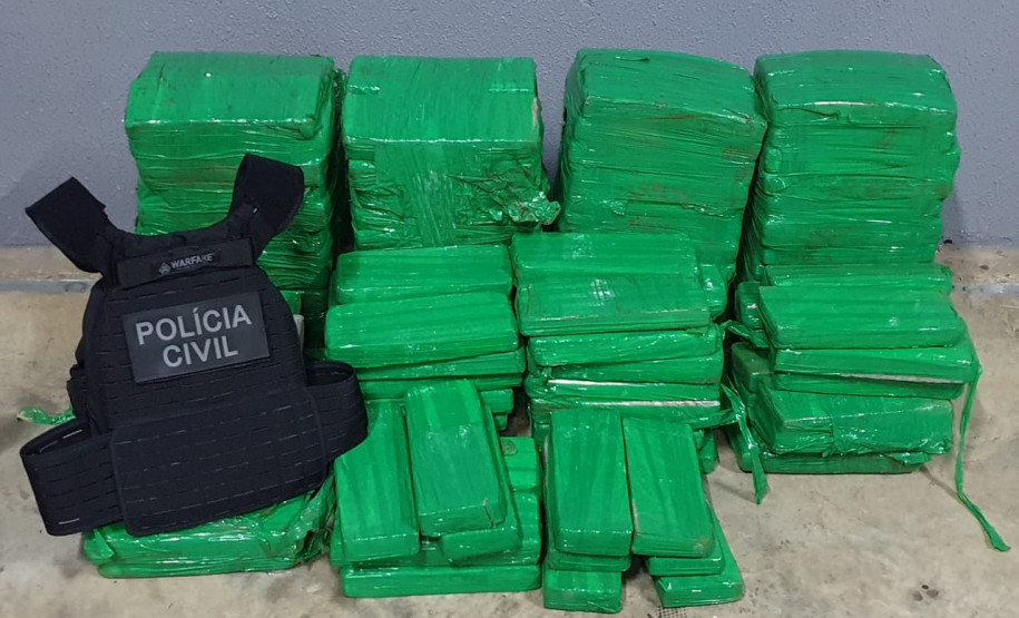 Diversos tabletes de droga agrupados e um colete da polícia civil, ao lado.