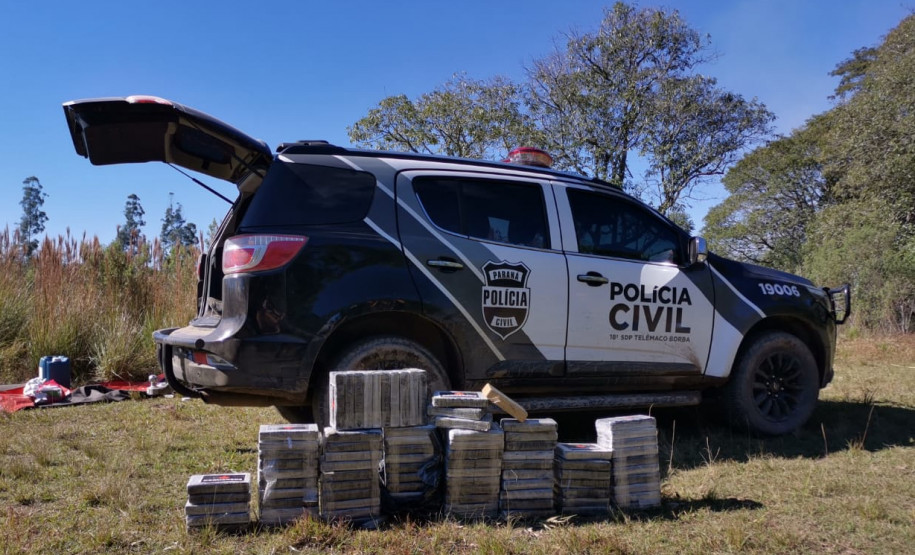 Diversos tabletes de droga empilhados ao lado de viatura da polícia civil