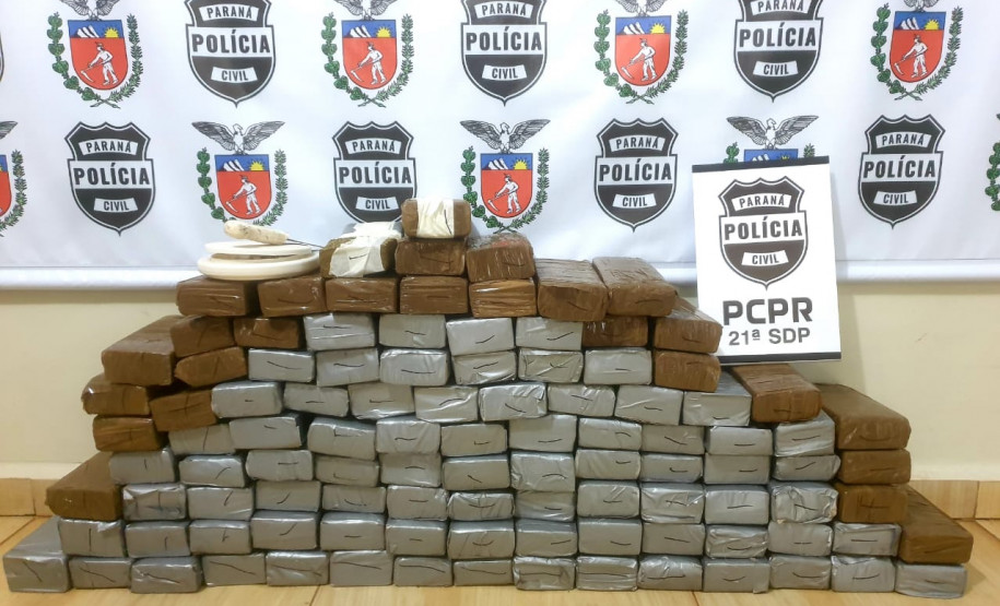 diversos tabletes da droga apreendida, empilhados no chão. Ao fundo, banner da polícia civil.