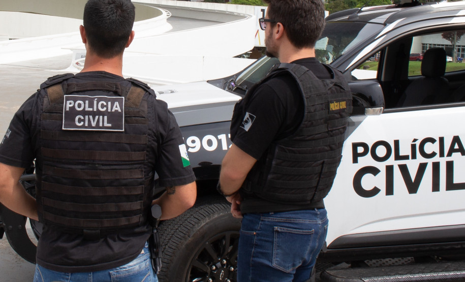 Dois policiais civis de costas, ao lado de viatura