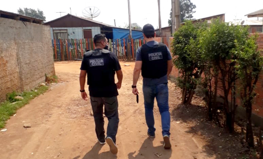 Dois policiais de costas, andando lado a lado, em rua de terra