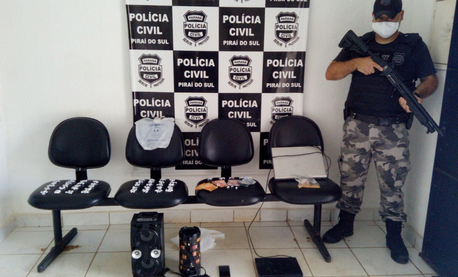 Policial civil empunhando arma ao lado de material apreendido. Ao fundo, banner da polícia civil.