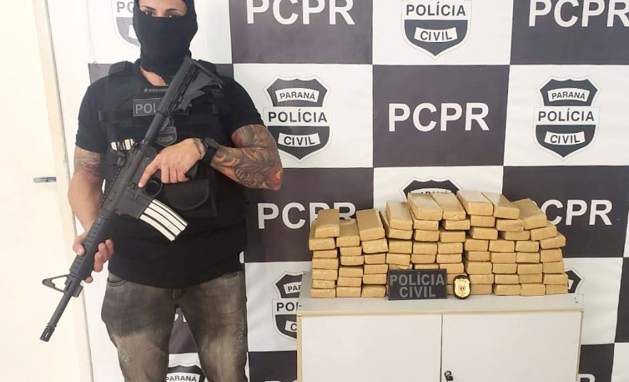 Policial empunhando arma ao lado de droga apreendida. ao fundo, banner da PCPR.