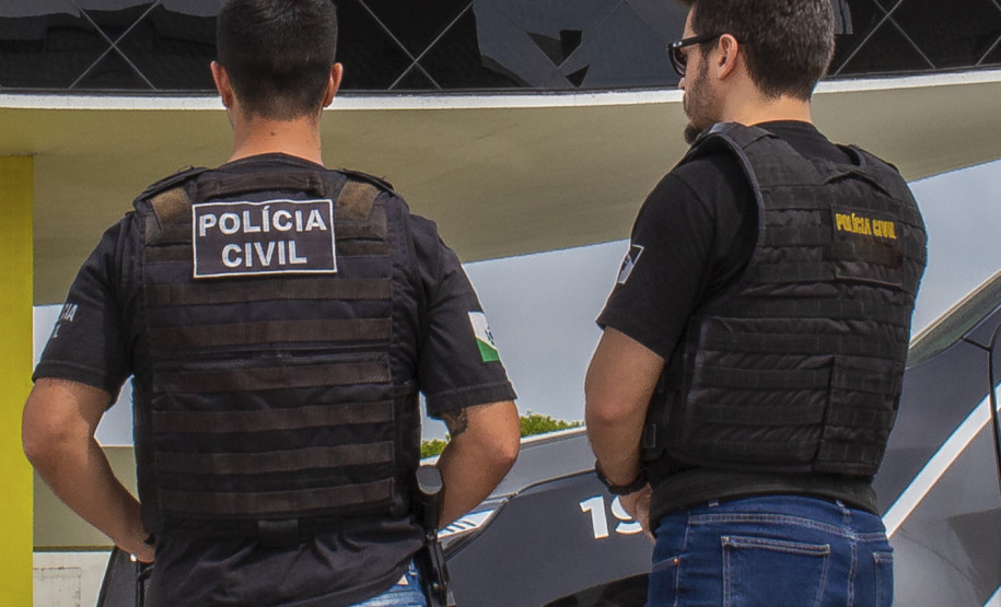 Dois policiais de costas, ao lado de viatura