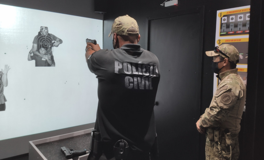 Policial aponta arma para alvo no simulador, observado pelo instrutor