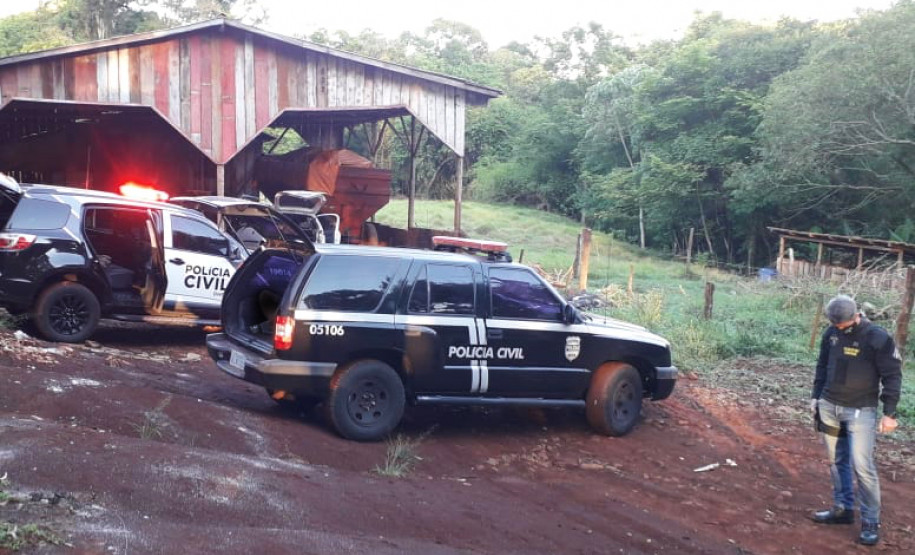 Policial civil olhando para baixo, ao lado de duas viaturas em área rural