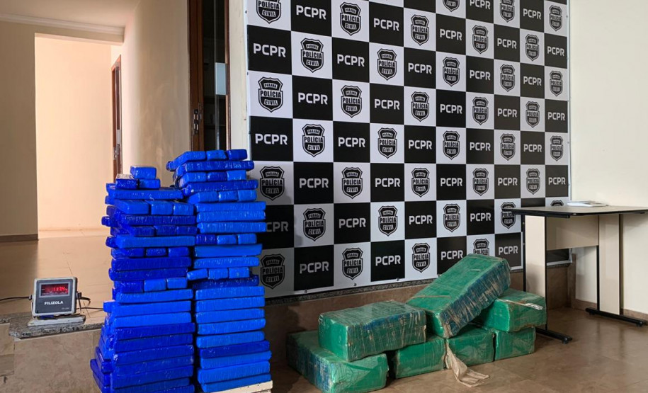 Tabletes de droga empilhados. ao fundo, banner com o brasão da polícia civil.