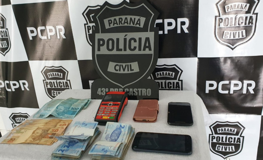 Material apreendido, sobre uma mesa. Ao fundo banner e logo da polícia civil