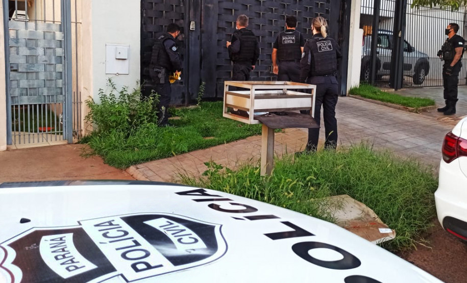 Policiais civis em frente a uma residência. Em primeiro plano, capô de viatura com brasão da polícia civil