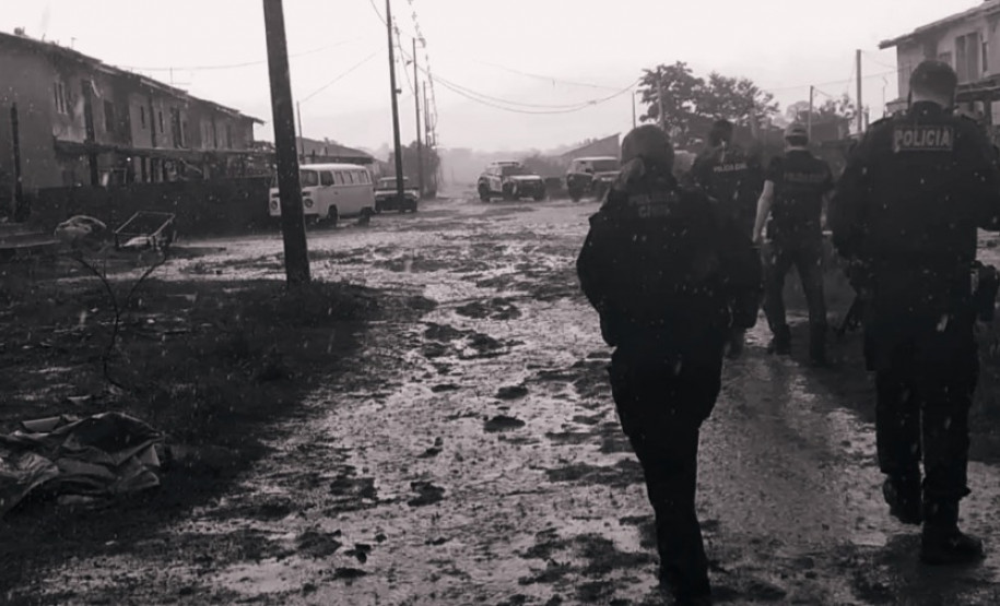 Policiais civis de costas, andando por via de terra após chuva