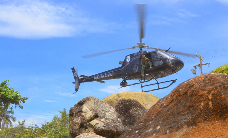 Helicóptero da policia civil levantando voo no litoral. Diversas rochas em primeiro plano.