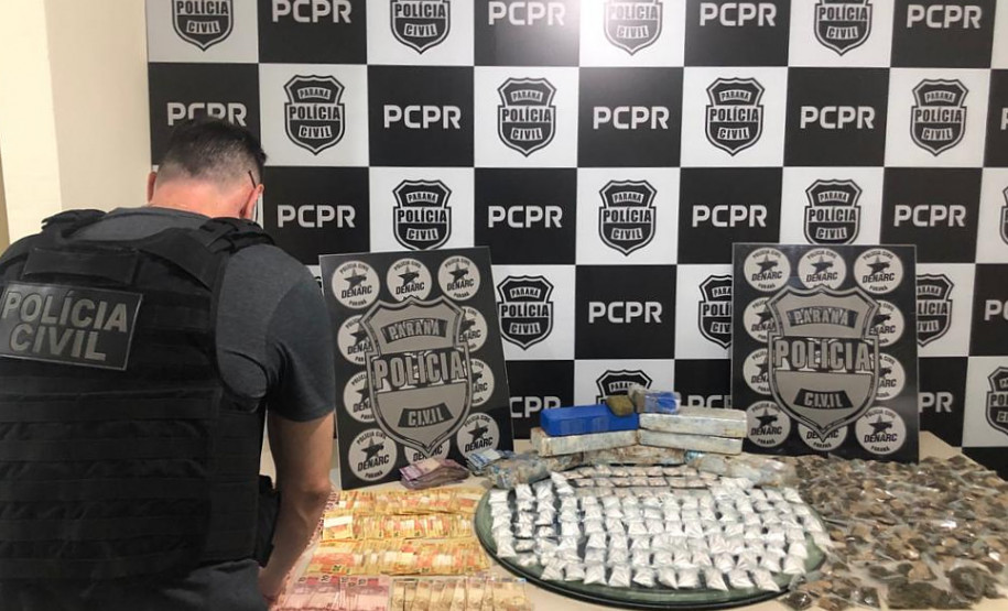 Policial civil de costas, organizando material apreendido sobre uma mesa. Ao fundo, banner com logo da PCPR.