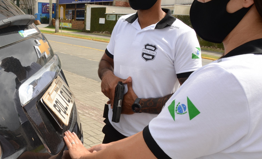 Policial civil fecha tampa do porta-malas de viatura enquanto outro policial empunha arma