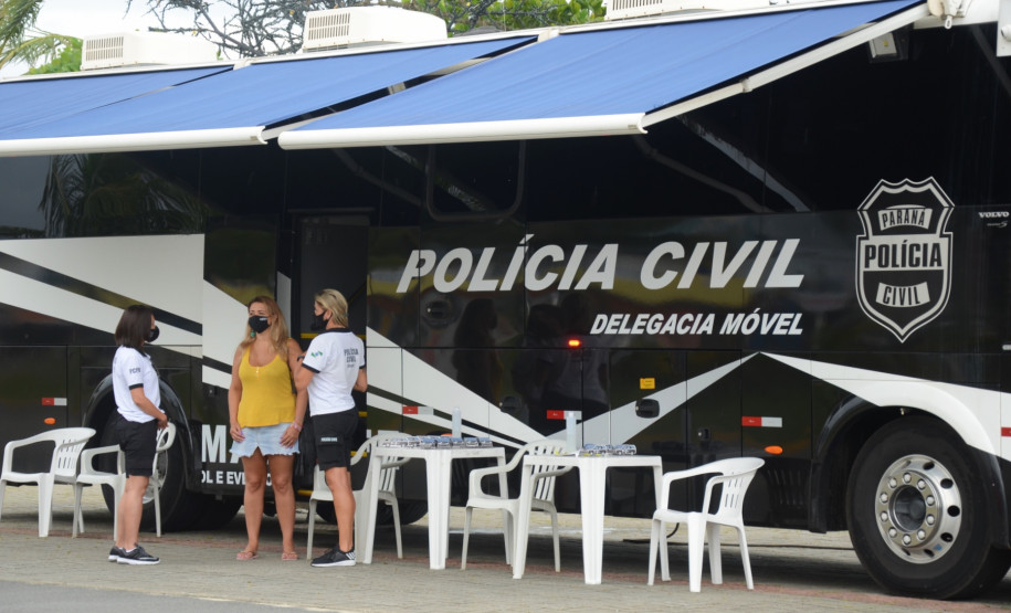 Duas policiais civis atendendo uma veranista ao lado do ônibus com a delegacia móvel