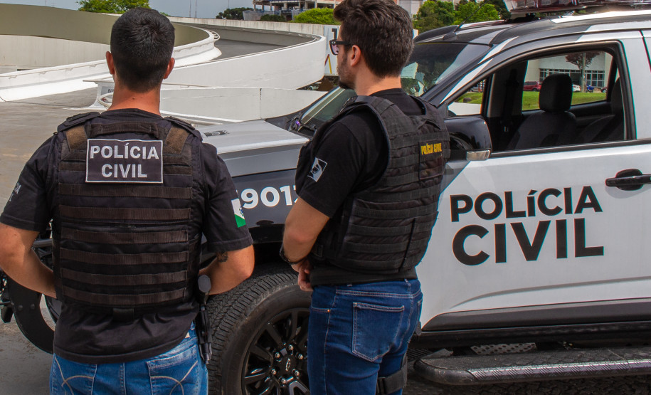 Dois policiais de costas, ao lado de viatura