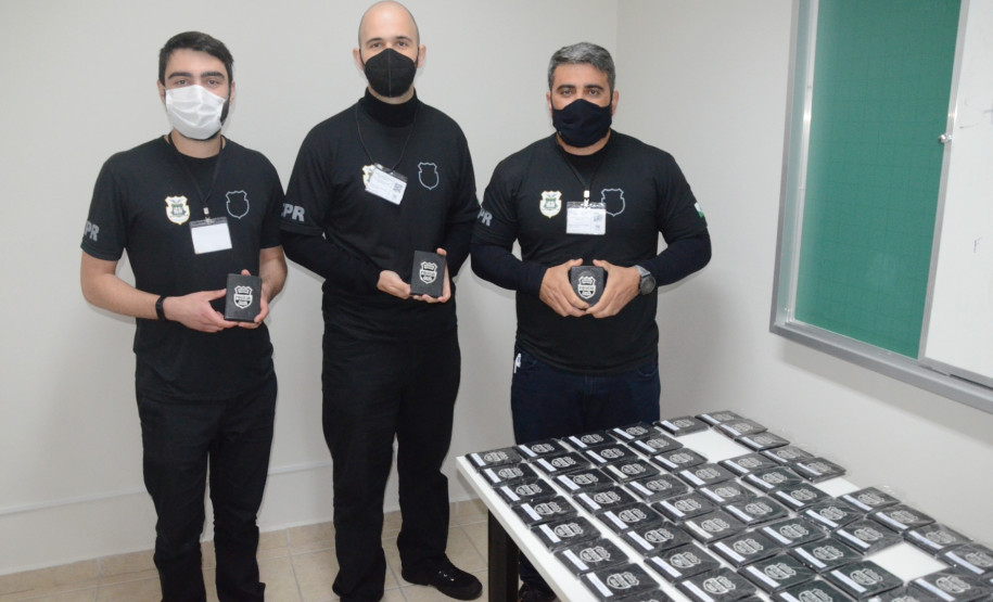 Três alunos mostrando carteira funcional recebida. Em primeiro plano, várias carteiras sobre uma mesa