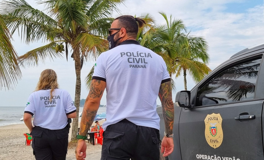Dois policiais civis ao lado de viatura estacionada à beira-mar