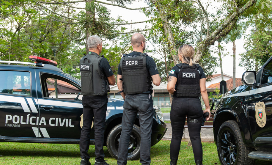 Três policiais civis de costas ao lado de viaturas