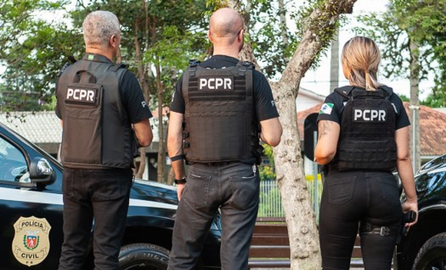 Três policiais de costas ao lado de viaturas