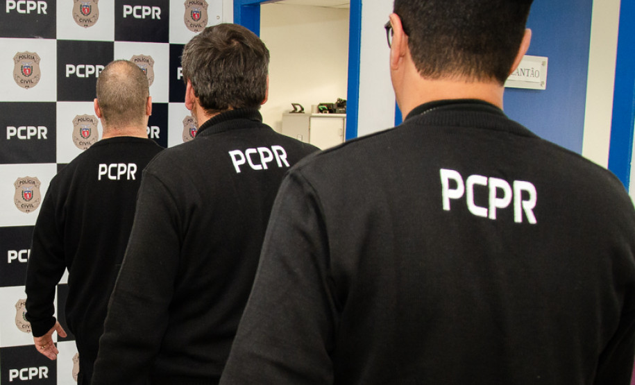 Três policiais civis de costas. Ao fundo, banner da PCPR