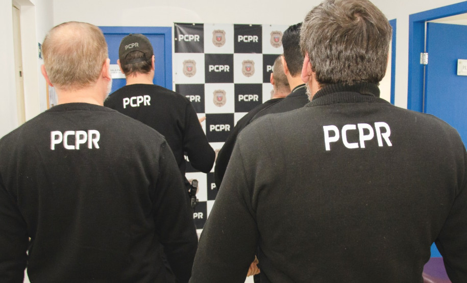 Vários policiais civis de costas. Ao fundo, banner da PCPR