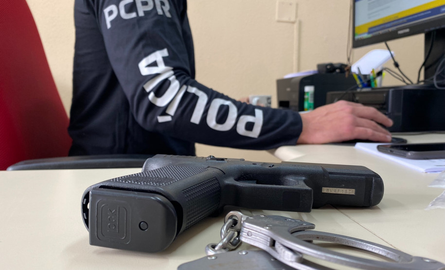 Policial civil ao computador. Em primeiro plano, arma e algemas