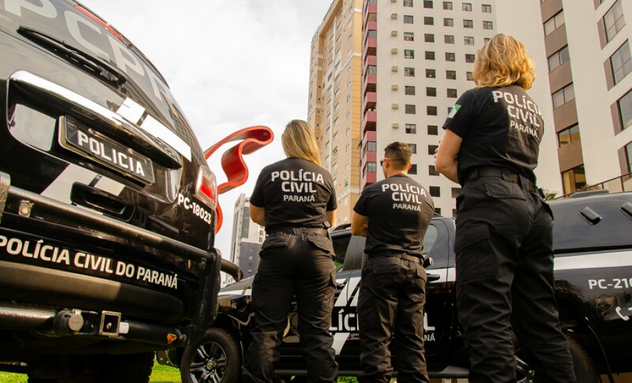 Três policiais civis de costas entre viaturas