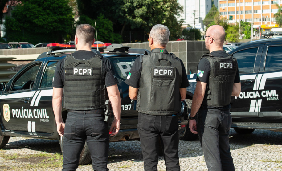 Três policiais civis ao lado de viaturas